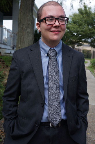 Silas stands outside on a sidewalk in a suit and paisley tie, hands in pockets, smiling